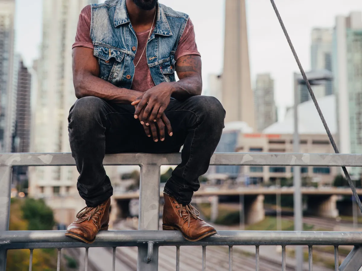 Homme assis sur une rambarde en ville, jean noir ajusté et bottines en cuir marron lacées