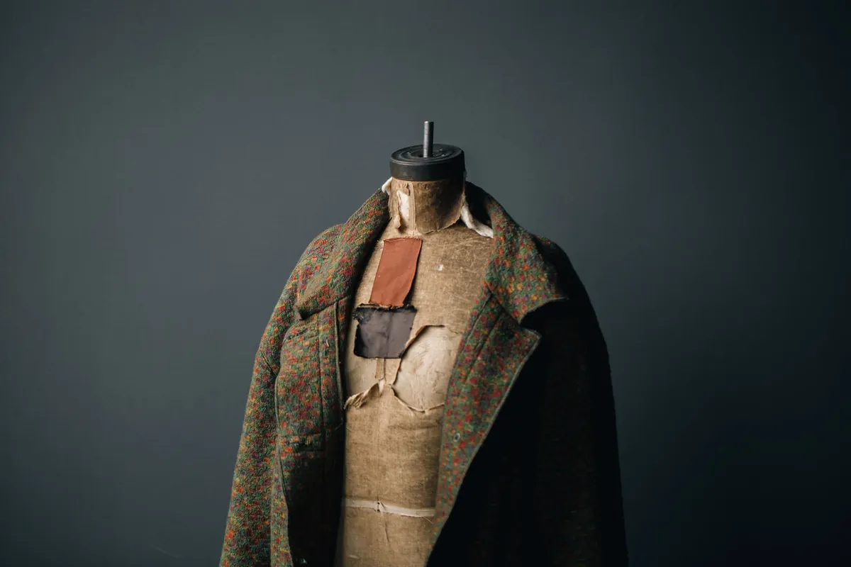 Manteau en drap de laine chiné vert et rouille posé sur un buste de couturier, devant un fond gris