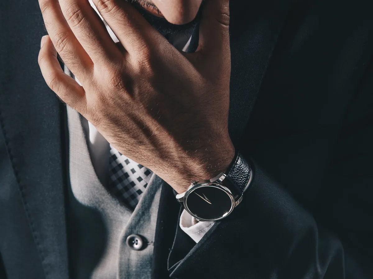 Man in a dark suit jacket over a buttoned grey vest, with a checked tie and a black-dial watch