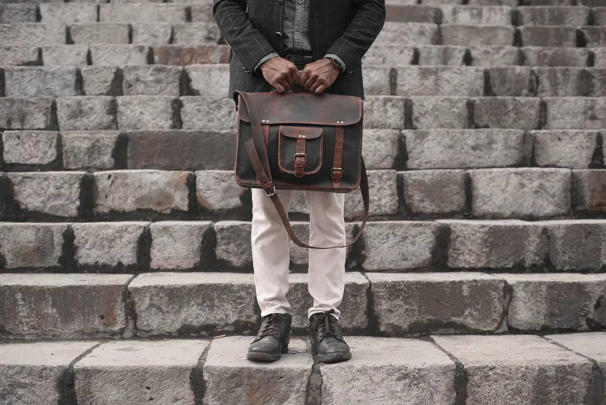 Man in a dark jacket and light trousers holding a brown leather bag in front of stone steps