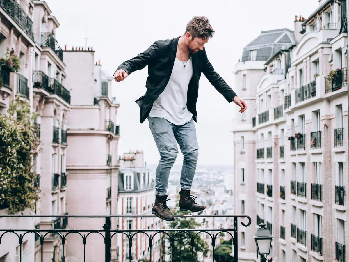 Man in a dark jacket, white T-shirt, faded jeans and brown boots, on a railing above the Paris rooftops