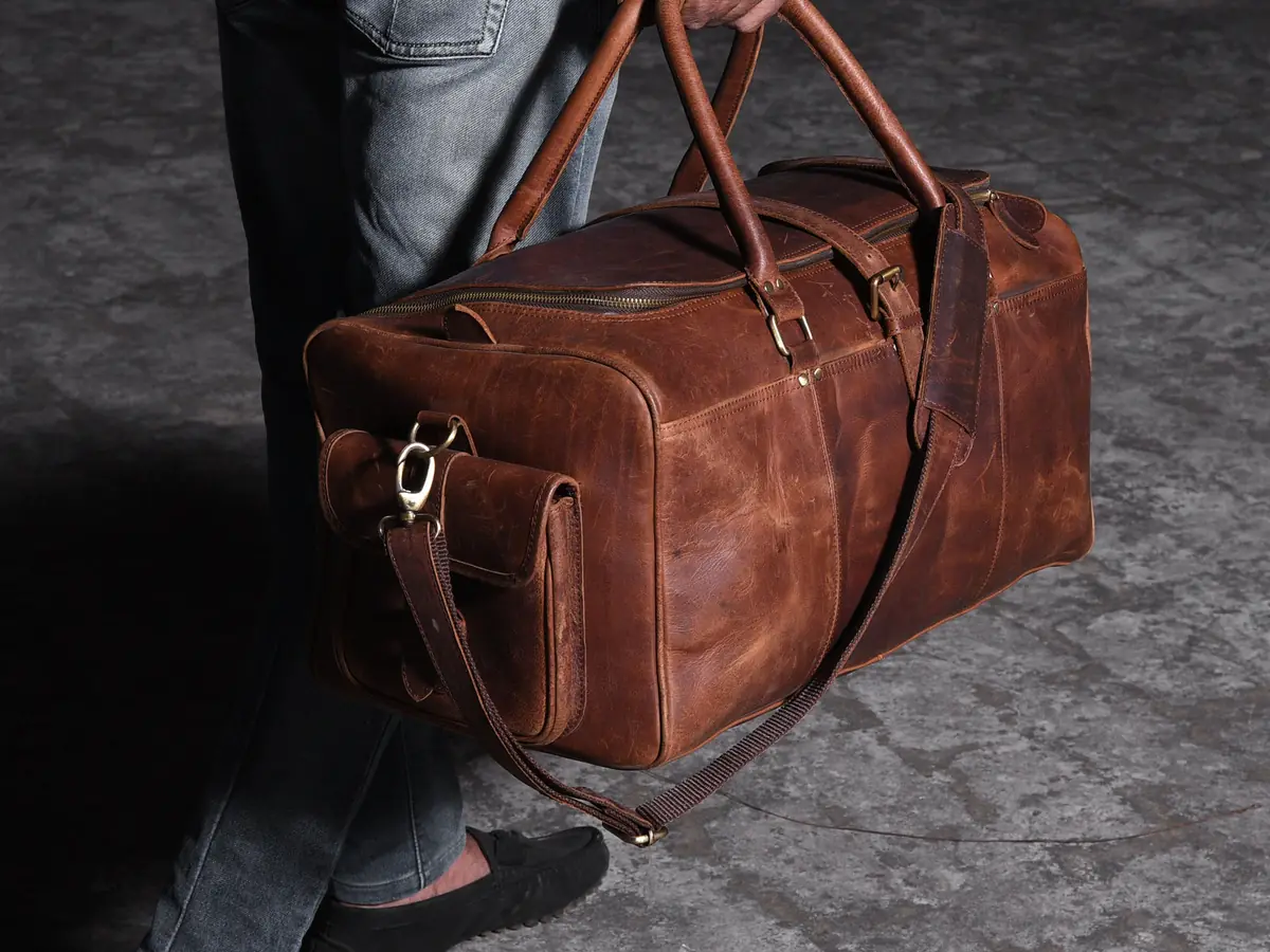 Homme en jean et mocassins portant à la main un sac week-end en cuir brun, sur un sol de pierre